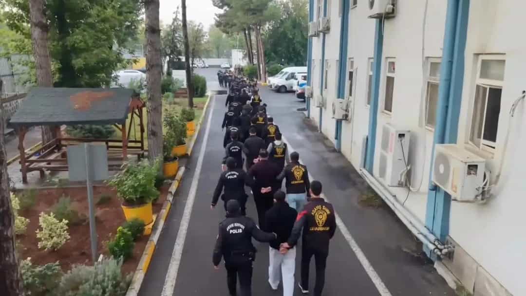 Polis ekipleri, gözaltına alınan şüphelileri bir bina önündeki yolda yürütüyor.