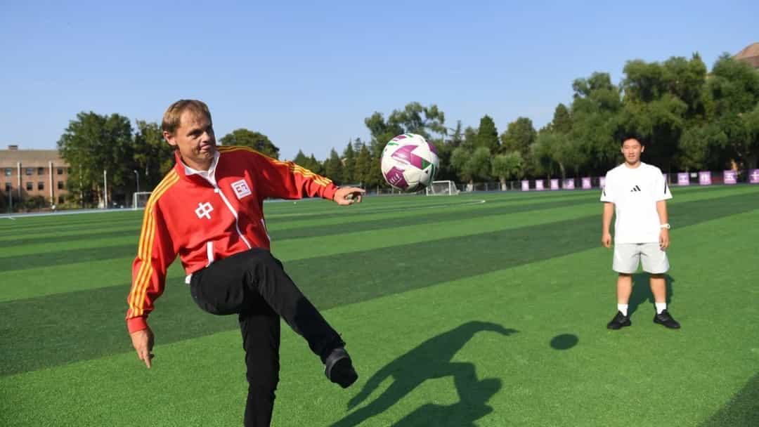 Bir futbol sahasında kırmızı ceketli bir adam havadaki topa vururken, beyaz formalı bir kişi onu izliyor.