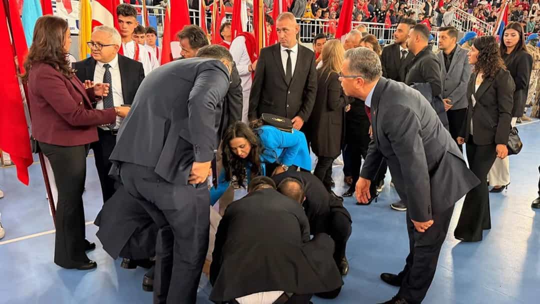 Tören sırasında yere yığılan öğrenciye çevresindekiler yardım ederken, gri takım elbiseli bir kişi müdahalede bulunuyor.