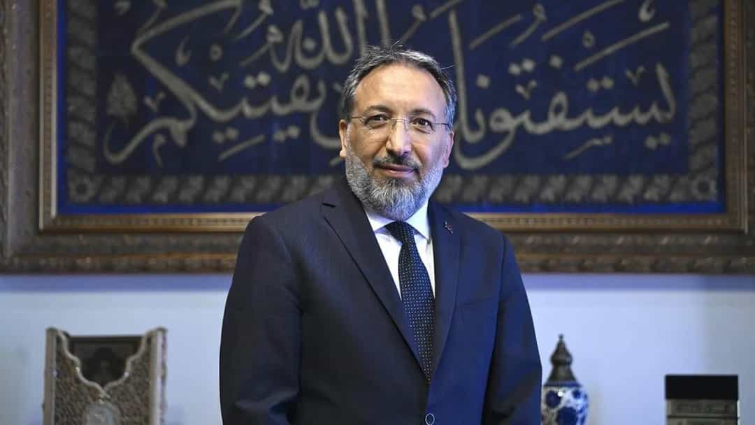 Diyanet İşleri Başkanı Safi Arpaguş, hat yazılı bir panonun önünde takım elbiseyle poz veriyor.