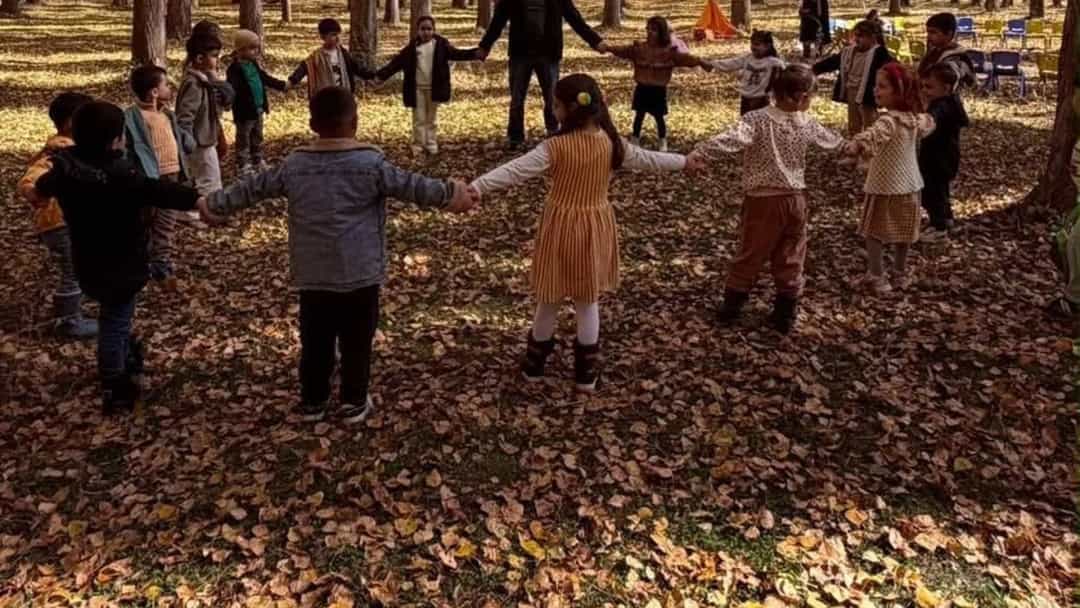 Çocuklar sonbahar yapraklarıyla kaplı bir ormanda el ele tutuşarak oyun oynuyor.