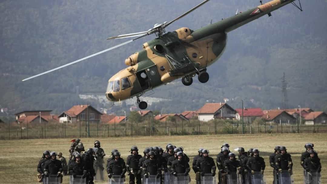 Kamuflajlı bir helikopter, güvenlik güçlerinin bulunduğu alanın üzerinde uçuyor.
