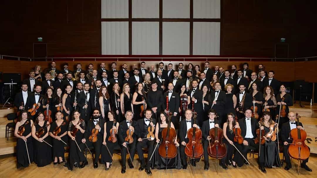 Bir konser salonunda yaylı çalgılarıyla birlikte poz veren büyük bir orkestra ve koro topluluğu.