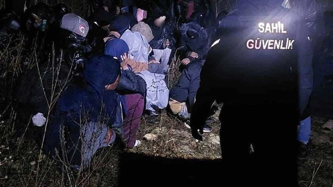 Gece saatlerinde sahil güvenlik görevlisinin önünde, arazide bir arada bulunan göçmenler.