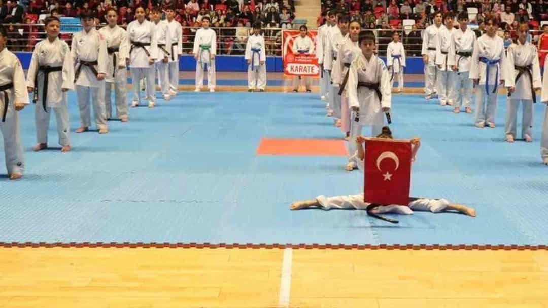Bir spor salonunda Türk bayrağı eşliğinde gösteri yapan sporcular, tribünlerdeki izleyicilerin önünde yer alıyor.