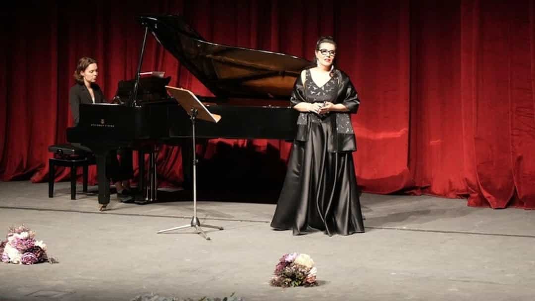 Kırmızı perdeli sahnede bir solist, piyano eşliğinde konser veriyor.