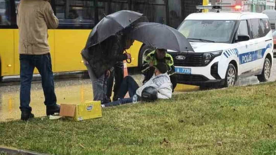 Yağmurlu havada yerdeki motosiklet sürücüsünü şemsiyeyle koruyan trafik polisi