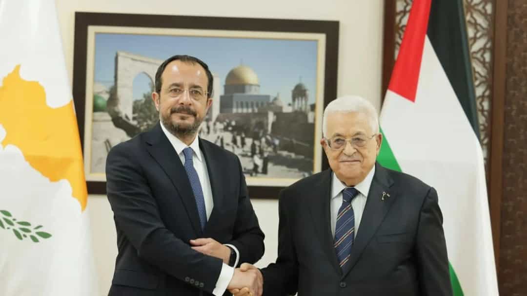 Mahmud Abbas, Filistin ve Kuzey Kıbrıs Türk Cumhuriyeti bayrakları önünde bir yetkiliyle tokalaşıyor.
