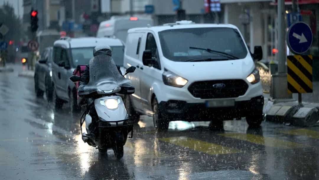 Yağmurlu ve su birikintileriyle kaplı yolda ilerleyen motosikletli, yanında seyreden beyaz bir minibüs.