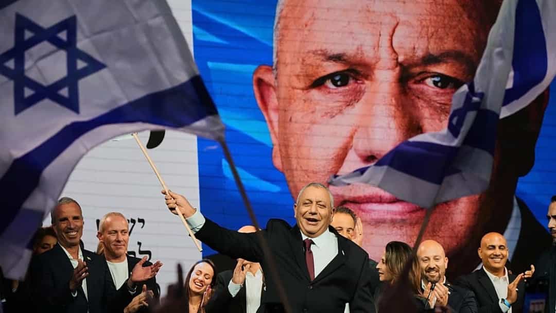 Binyamin Netanyahu, İsrail bayrağını kaldırarak bir siyasi etkinlikte konuşurken arkasında büyük bir portresi görülüyor.