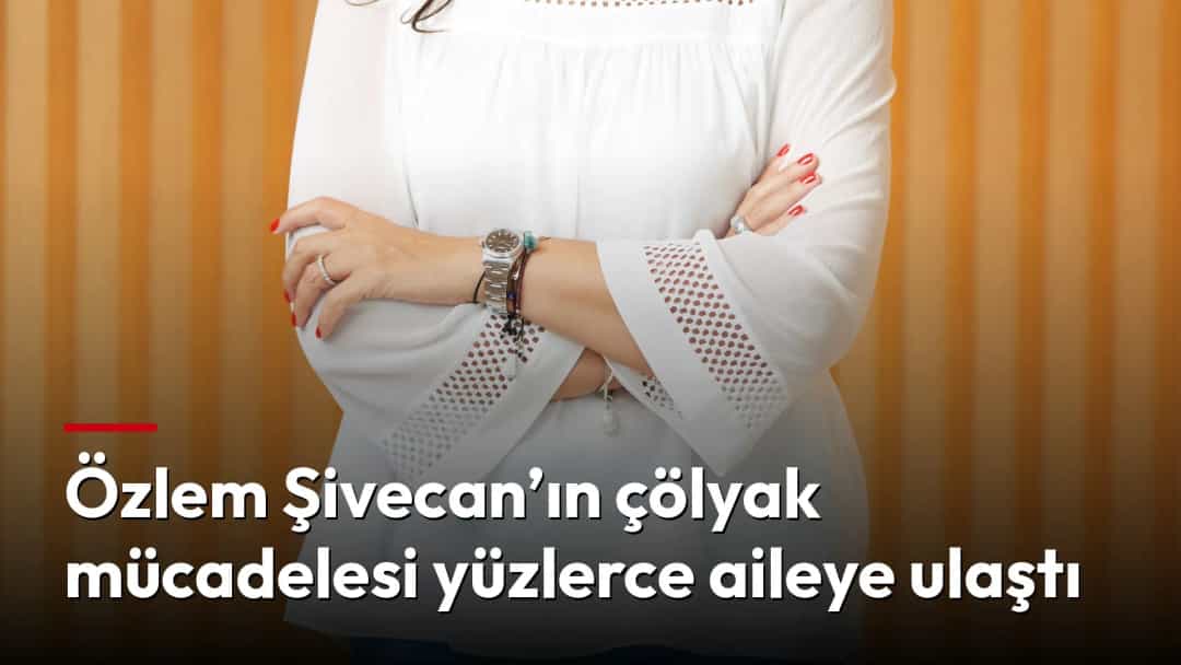 Özlem Şivecan, beyaz bluz ve mavi pantolonla, ahşap çıtalı bir duvarın önünde poz veriyor.