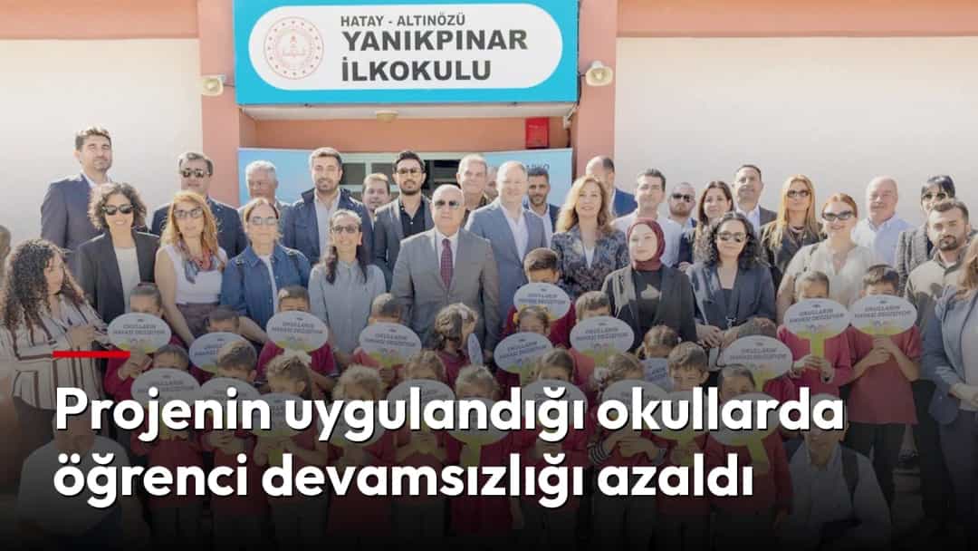Öğrenciler ve yetişkinler, okul girişinde toplu halde duruyor; ön sıradaki çocuklar yazılı kartlar tutuyor.