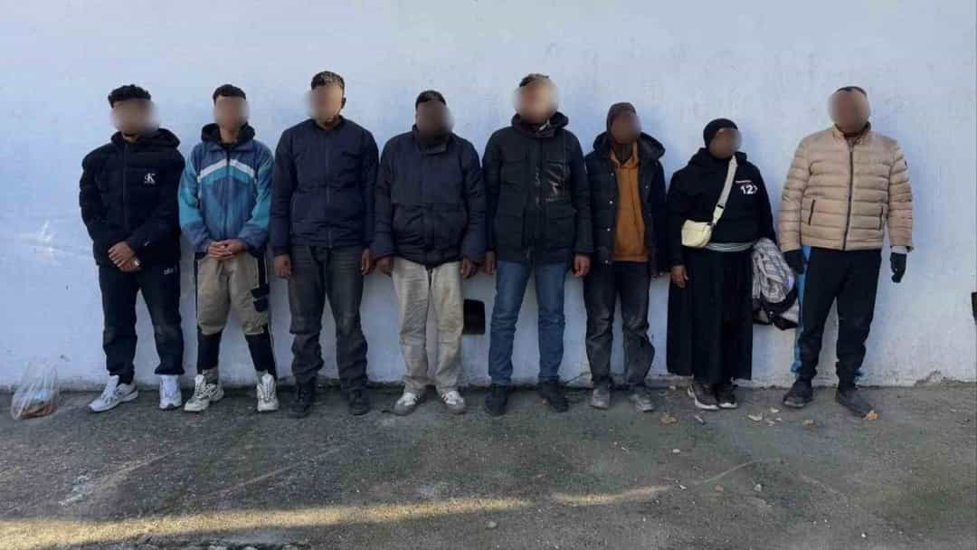 Yakalanan düzensiz göçmenler bir duvarın önünde yan yana duruyor.